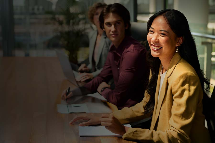 Smiling business professional participates in a collaborative meeting while seated at a conference table with colleagues in a modern office. A laptop, notebook, and natural light create a welcoming, professional learning environment.