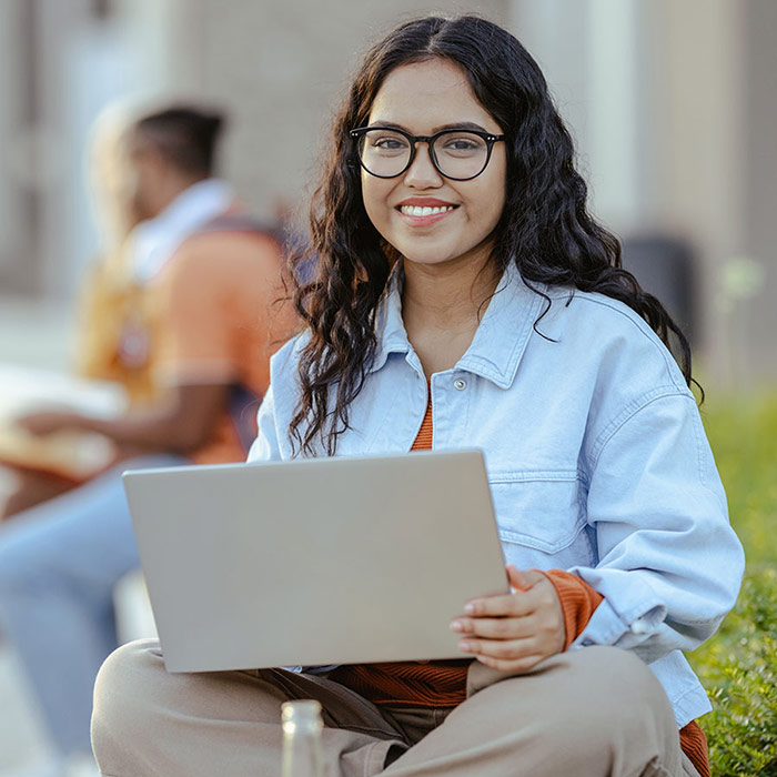 Student with glasses sits outside using a laptop, smiling at the camera with a campus setting in the background.