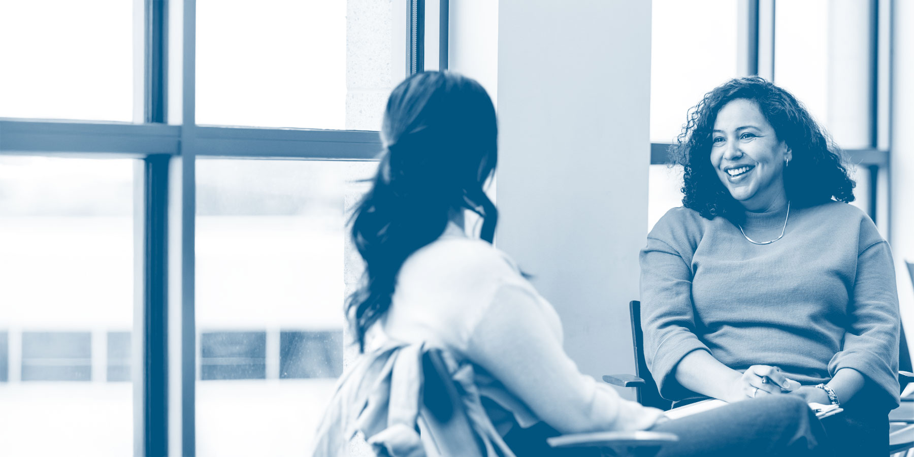 Two women sit facing each other in a bright room, smiling and engaged in conversation during a counseling or advising session.