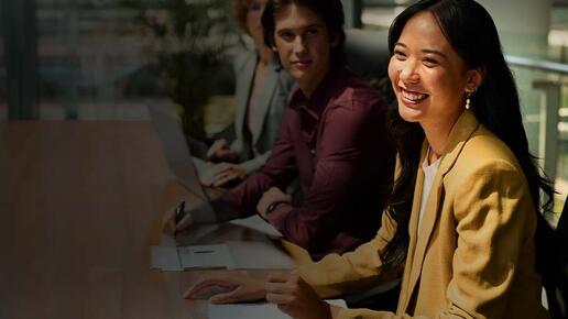 Smiling business professional participates in a collaborative meeting while seated at a conference table with colleagues in a modern office. A laptop, notebook, and natural light create a welcoming, professional learning environment.