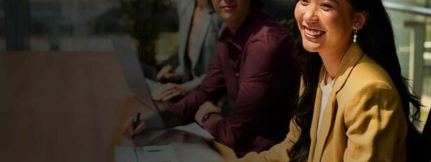 Smiling business professional participates in a collaborative meeting while seated at a conference table with colleagues in a modern office. A laptop, notebook, and natural light create a welcoming, professional learning environment.