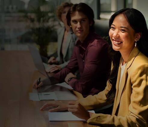 Smiling business professional participates in a collaborative meeting while seated at a conference table with colleagues in a modern office. A laptop, notebook, and natural light create a welcoming, professional learning environment.