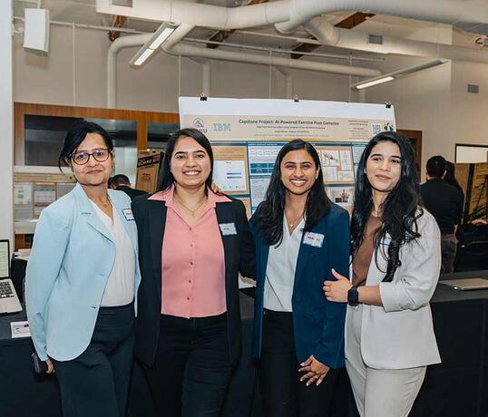 Four students in business attire stand smiling in front of a research poster during a university capstone project showcase. The poster, titled "AI-Powered Exercise Pose Correction," is displayed in an indoor exhibition space with other student projects visible in the background.