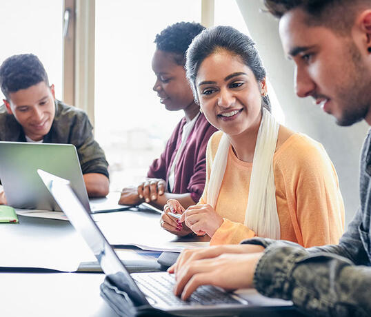 A group of diverse students sit together at a table, collaborating on laptops and smiling during a study session.