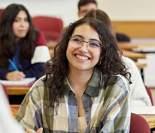 Smiling student with glasses sits in a classroom, holding a pen and looking toward the camera while classmates study in the background.