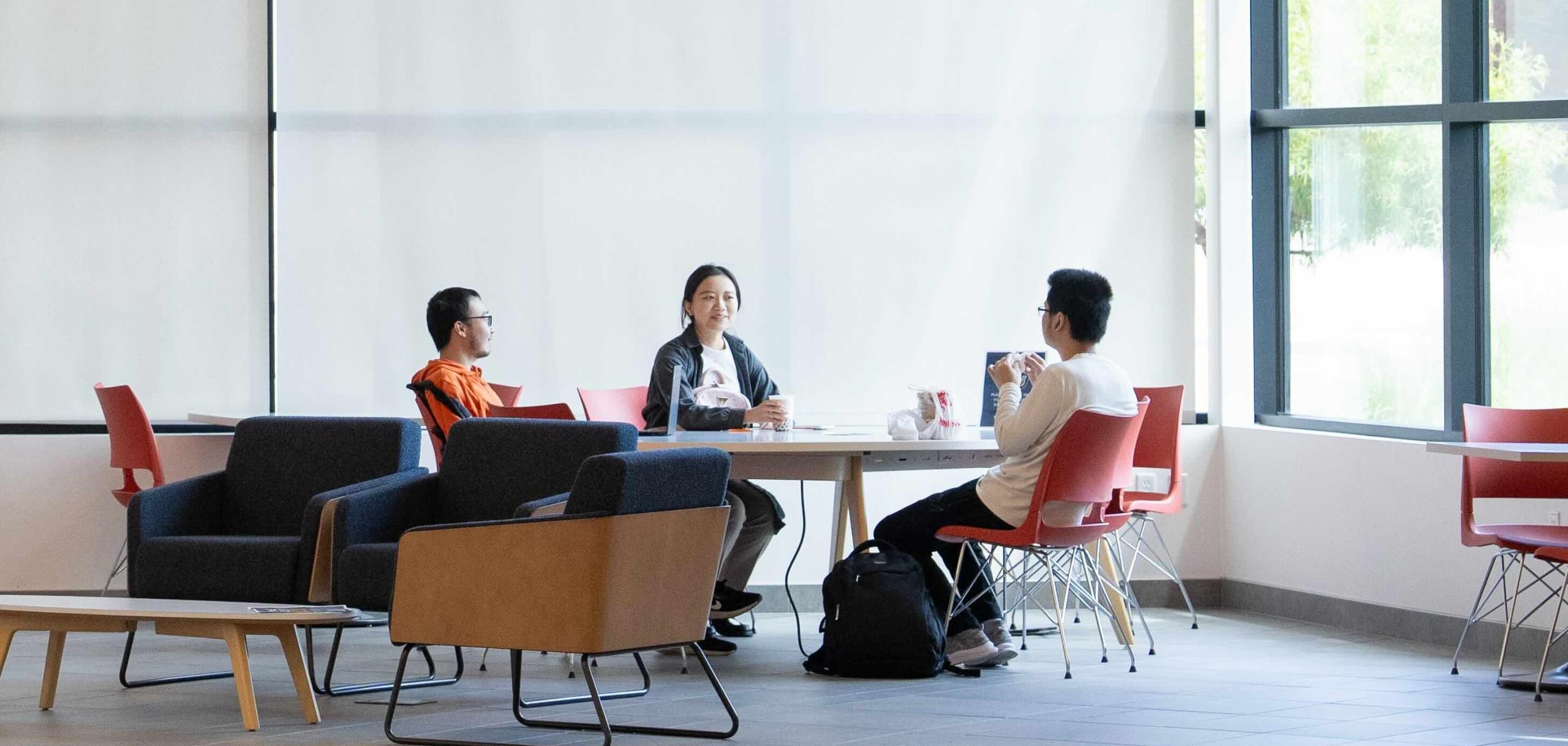 Three students are casually chatting in the lobby of the SFBU campus. The lobby is modern and welcoming, with comfortable seating areas and natural light filtering through large windows. 