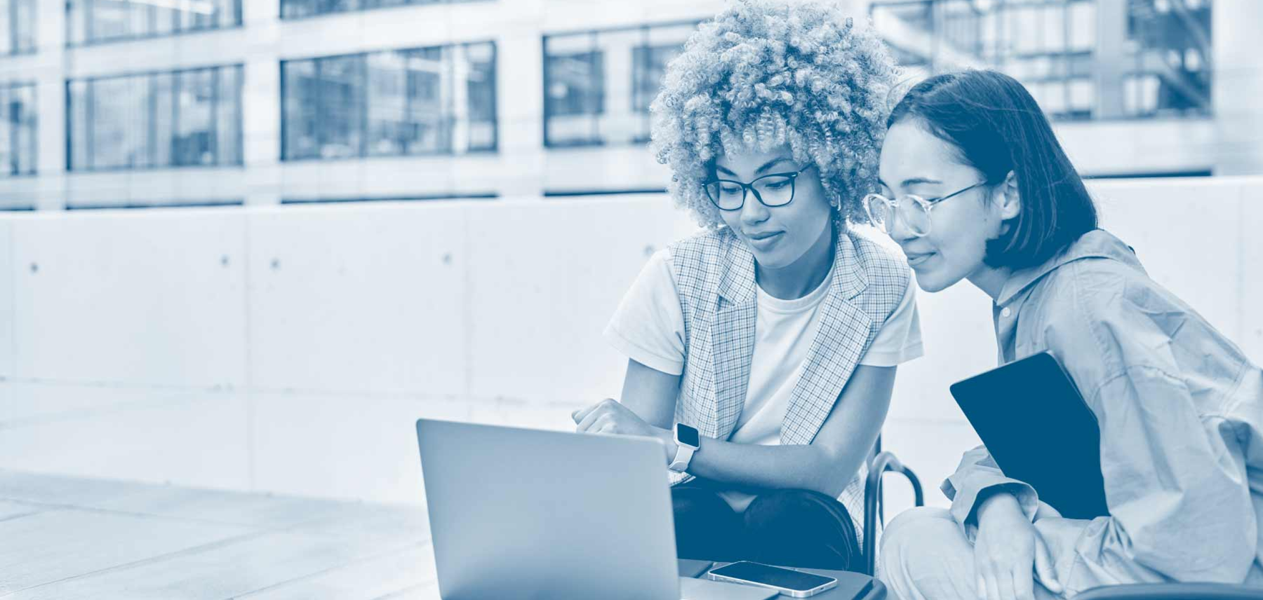 Two students sit together outdoors, looking at a laptop while collaborating on coursework. One student points at the screen, and the other holds a tablet. The image features a blue monochrome overlay and a modern campus building in the background, representing teamwork and graduate business education.