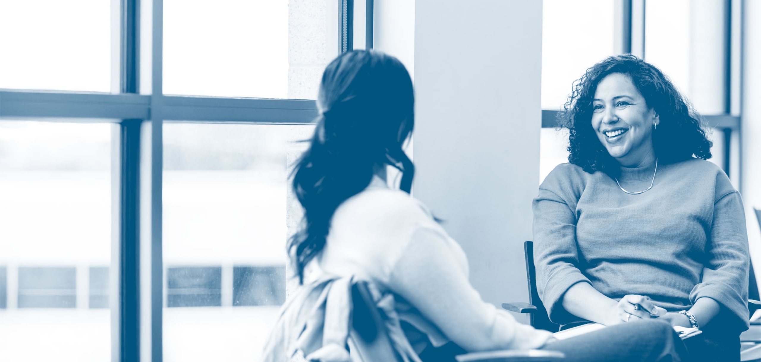 Two women sit facing each other in a bright room, smiling and engaged in conversation during a counseling or advising session.
