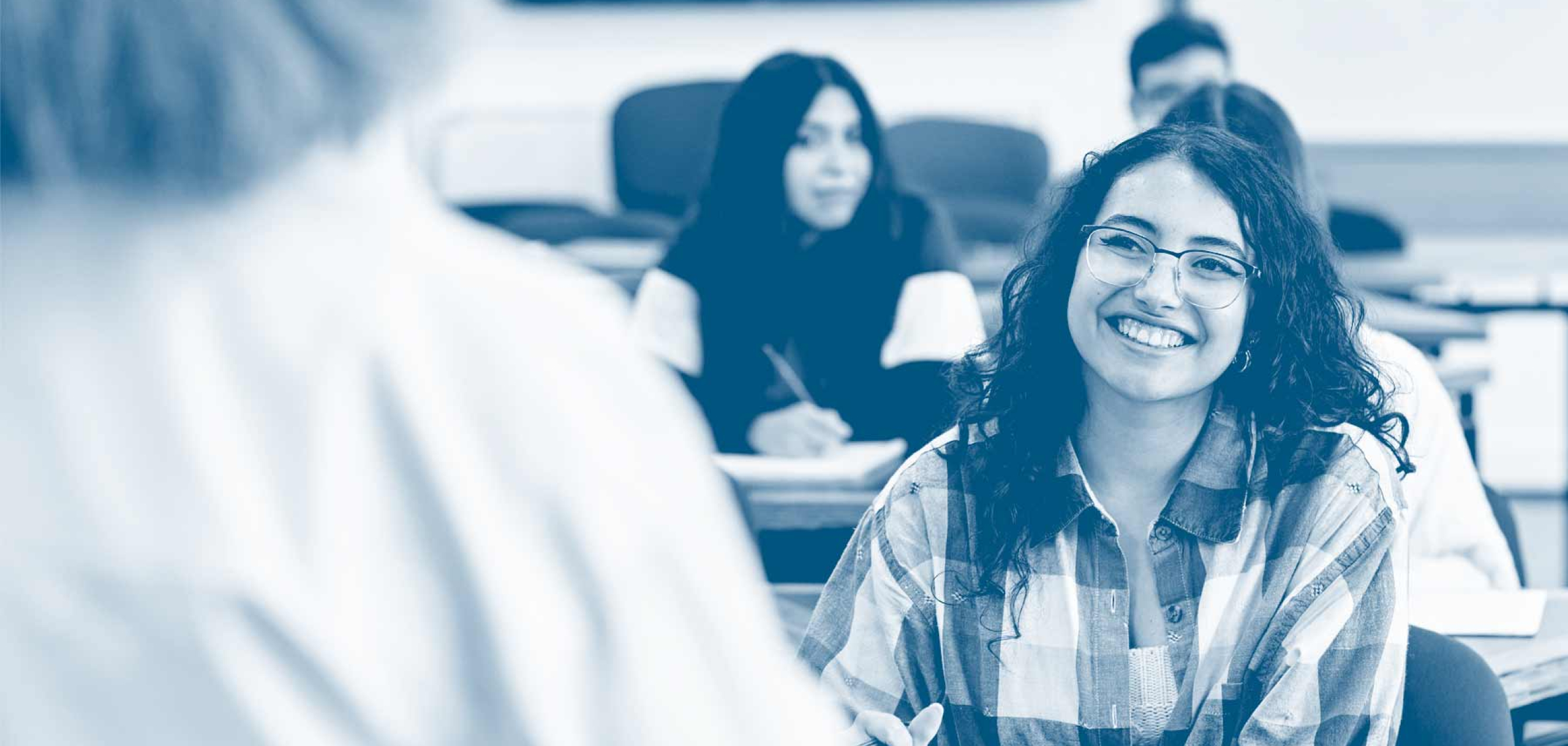 Smiling student with glasses sits in a classroom, engaging with an instructor while classmates take notes in the background.