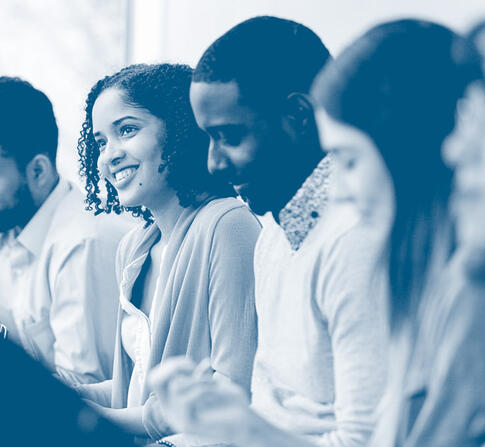 A group of students sit side by side in a classroom, smiling and reviewing notes, with a focus on a student in the center engaged in discussion.