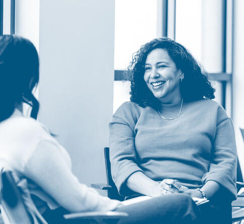 Two women sit facing each other in a bright room, smiling and engaged in conversation during a counseling or advising session.