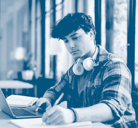 Student wearing headphones studies at a laptop in a modern campus workspace, taking notes while seated at a table.