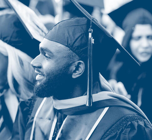 Graduates wearing caps and gowns sit together during a commencement ceremony, smiling and looking ahead, with a focus on a bearded graduate in the foreground.