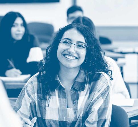 Smiling student with glasses sits in a classroom, engaging with an instructor while classmates take notes in the background.