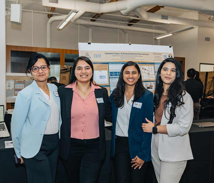 Four students in business attire stand smiling in front of a research poster during a university capstone project showcase. The poster, titled "AI-Powered Exercise Pose Correction," is displayed in an indoor exhibition space with other student projects visible in the background.