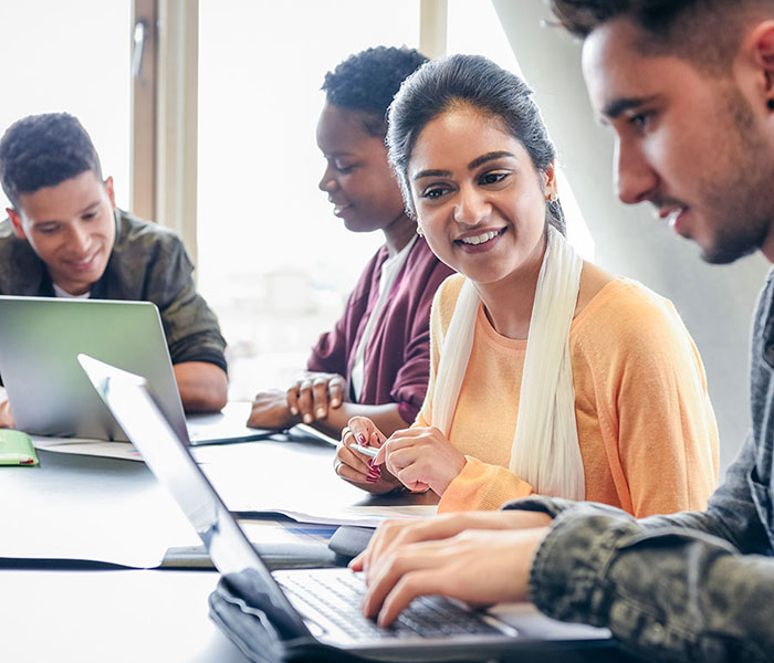 A group of diverse students sit together at a table, collaborating on laptops and smiling during a study session.