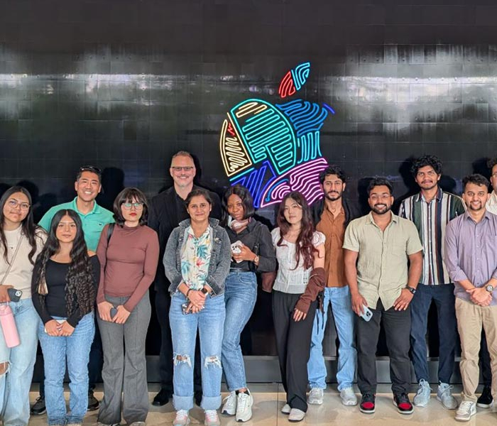Group of students and an instructor stand together in front of a wall with a colorful Apple logo, posing for a group photo.