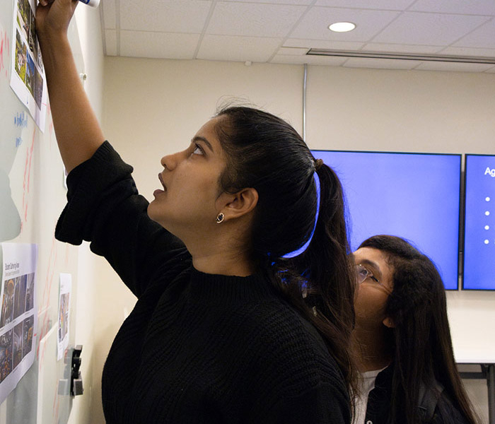 Student writes on a whiteboard during a collaborative classroom activity while another student observes.