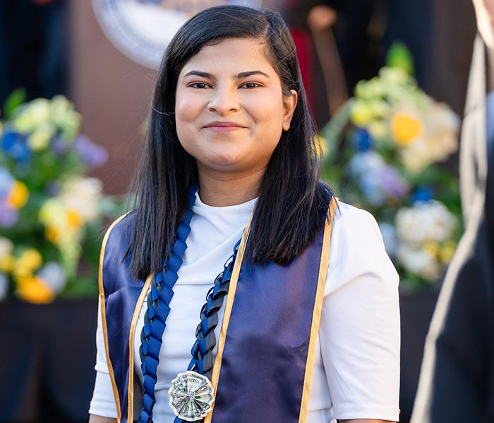 Graduate wearing a stole and medals smiles during an outdoor commencement ceremony.