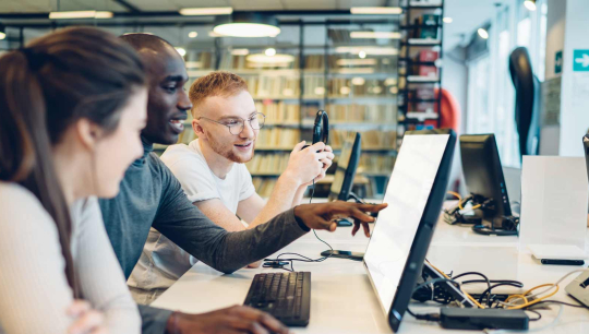 Students collaborate at desktop computers in a lab, discussing work and pointing at a screen.