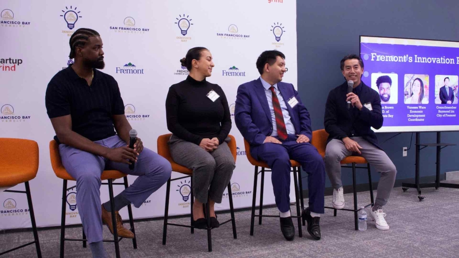 Four panelists sit on stage during Fremont’s Innovation Forum, holding microphones and engaging in discussion. A branded backdrop and presentation screen with panelist names and photos are behind them.