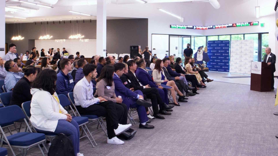 A large audience seated in rows listens attentively to a speaker at a podium during Fremont’s Innovation Forum. The event takes place in a modern space with bright lighting and a stock ticker display in the background.