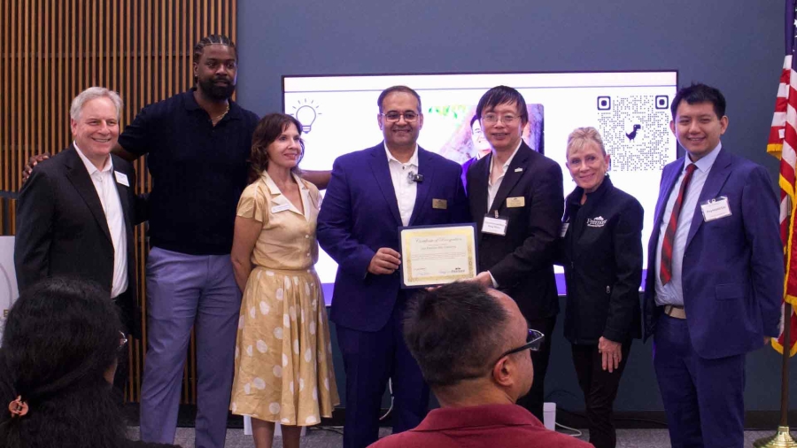 A group of seven people pose for a photo at Fremont’s Innovation Forum, with one man holding a certificate of recognition. Smiling participants stand in front of a presentation screen and U.S. flag.